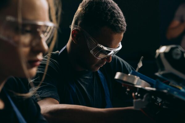 Two apprentices working intently in an industrial setting, wearing safety goggles. The focus is on a young man in the foreground who is concentrating on a piece of machinery, with a young woman slightly out of focus in the background also looking at the equipment.