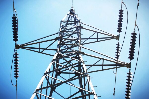 Alt text: "A close-up view of an electricity pylon against a clear blue sky. The steel lattice structure is prominently displayed, with several sets of insulators hanging from the cross arms, connecting to overhead power lines.