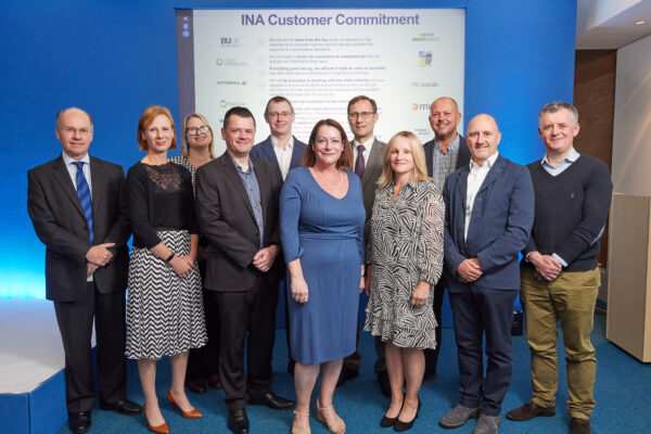 Group photo of eleven senior leaders of the Independent Networks Association (INA), standing in front of a presentation screen displaying the INA Customer Commitment. They are dressed in business attire, with five individuals in the front row and six in the back, posing with a mix of smiles and neutral expressions. The backdrop is a bright blue wall with various company logos displayed alongside the commitments.