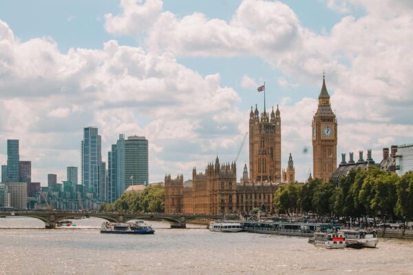 Panoramic view of the London skyline featuring the Houses of Parliament and the iconic Big Ben clock tower on the right, with the River Thames in the foreground where several boats are visible. Modern skyscrapers stand in the distance against a backdrop of fluffy white clouds in a blue sky, displaying a contrast between historical and contemporary architecture.
