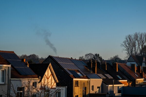 ooftops of a residential area during sunset, with solar panels visible on some of the houses, contrasting with a chimney emitting smoke in the background, symbolizing a mix of renewable energy adoption and traditional energy use.