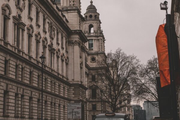 A photograph of the UK Treasury building in London. The historic, classical-style architecture features ornate stone detailing and prominent towers. A black taxi cab is driving along the street in front of the building, and bare trees line the pavement. The sky is overcast, giving the scene a muted, grey tone.