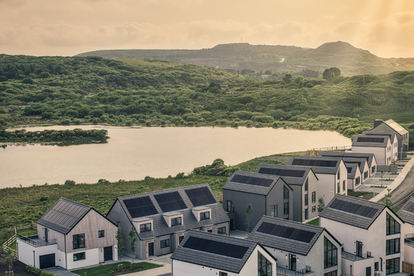 Aerial view of modern eco-friendly homes with solar panels at West Carclaze Garden Village, set beside a lake and surrounded by green hills at sunset.